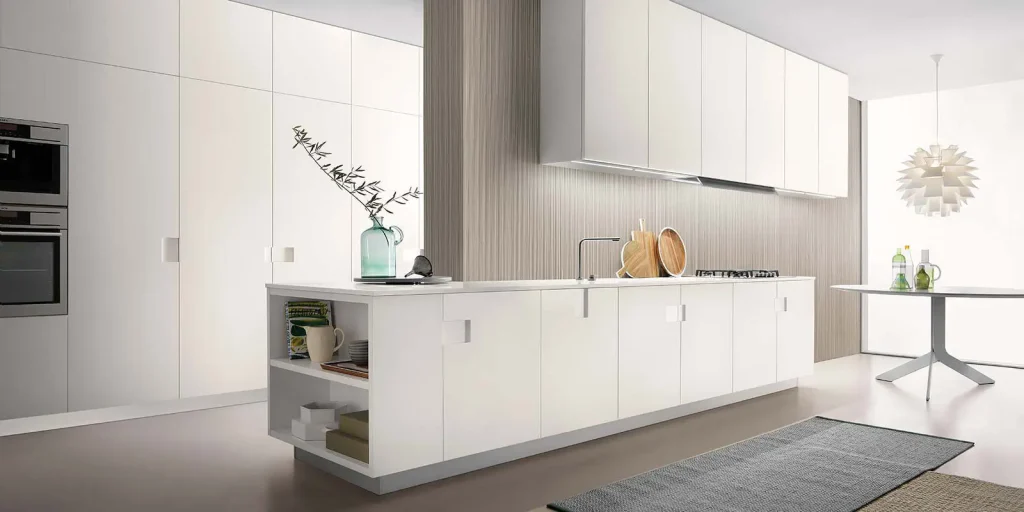 This photo shows a stylish and modern living room with a cozy and refreshing atmosphere. The room features a light gray sectional sofa with pastel green cushions and a matching ottoman placed on a soft white rug. Behind the sofa, the accent wall is painted in a calming green shade, decorated with unique floating cube shelves holding books and small decor pieces. A tall floor lamp stands on one side, while on the opposite side, a white cabinet supports a television. Large windows with green curtains allow natural light to brighten the space, and a wicker chair with a plant beside it adds a natural, earthy touch. The combination of soft colors, greenery, and minimalistic furniture creates a warm, inviting, and contemporary look.